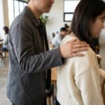 Medium shot of a man's hand on a woman's shoulder in a modern cafe, faces blurred. The gesture is subtle and ambiguous.
