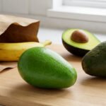 Three avocados on a wood board showing ripening stages: unripe, ripe, and cut open with pit. Banana in bag for ripening.