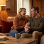 A couple shares a joyful laugh on a cozy brown leather sofa in a warm living room, illuminated by a fireplace and lamp.