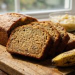 Golden-brown banana bread, sliced to show moist crumb, on a wood board with a ripe banana. Mashed bananas and flour in background. Natural light.