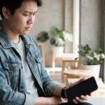 A man in a denim jacket looks thoughtfully at his smartphone's blank screen in a sunlit cafe, conveying introspection.
