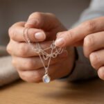 Close-up: Hands gently untangling a silver necklace with a luminous pendant, set against a soft, blurred background.