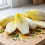 Fresh, crisp endives, whole and sliced to show pale yellow and white interior, on a wooden board with olive oil and herbs.