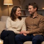 A couple in smart casual attire, smiling and holding hands on a couch in a softly lit living room, conveying love and security.