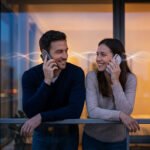 A man and woman on a balcony at dusk, holding smartphones to their ears and smiling warmly at each other, with light trails connecting their phones.