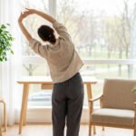 Woman in casual wear stretching by a large window in a modern home office with natural light, plants, and park view.