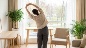 Woman in casual wear stretching by a large window in a modern home office with natural light, plants, and park view.