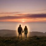Un couple vu de dos se tient la main, contemplant un magnifique coucher de soleil sur l'océan, entouré d'une aura lumineuse.