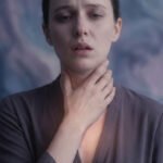 Gros plan sur une femme anxieuse, main sur la gorge. Son visage exprime la détresse, un cri étouffé. Lumière subtile sur la poitrine.
