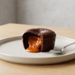 Close-up of a protein chocolate fondant with a flowing molten orange-brown core on a minimalist plate with a silver spoon.