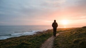 Silhouette marchant sur un sentier côtier herbeux, face à un coucher de soleil orange et rose sur l'océan calme.