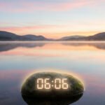 Lever de soleil pastel sur un lac brumeux, montagnes en arrière-plan. Un rocher avec "06:06" lumineux est au premier plan.