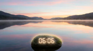 Lever de soleil pastel sur un lac brumeux, montagnes en arrière-plan. Un rocher avec "06:06" lumineux est au premier plan.